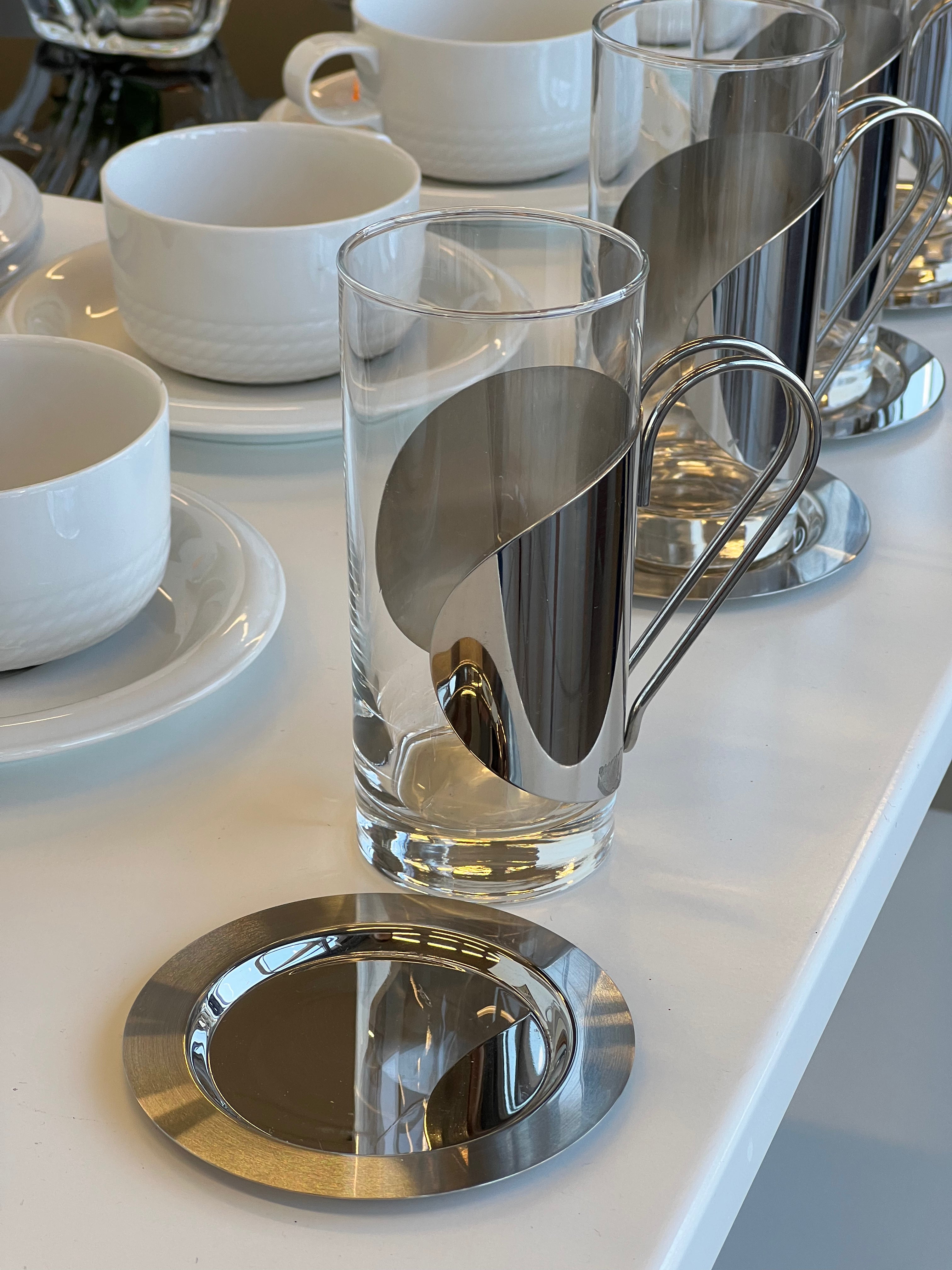 Nuotraukoje pavaizduotas stiklinis puodelis su metaliniu laikikliu, kuris apgaubia puodelį ir turi rankeną. Tokie puodeliai dažnai naudojami karštiems gėrimams, pavyzdžiui, arbatai ar kavai, patiekti, o metalinis laikiklis padeda apsaugoti rankas nuo karščio. 